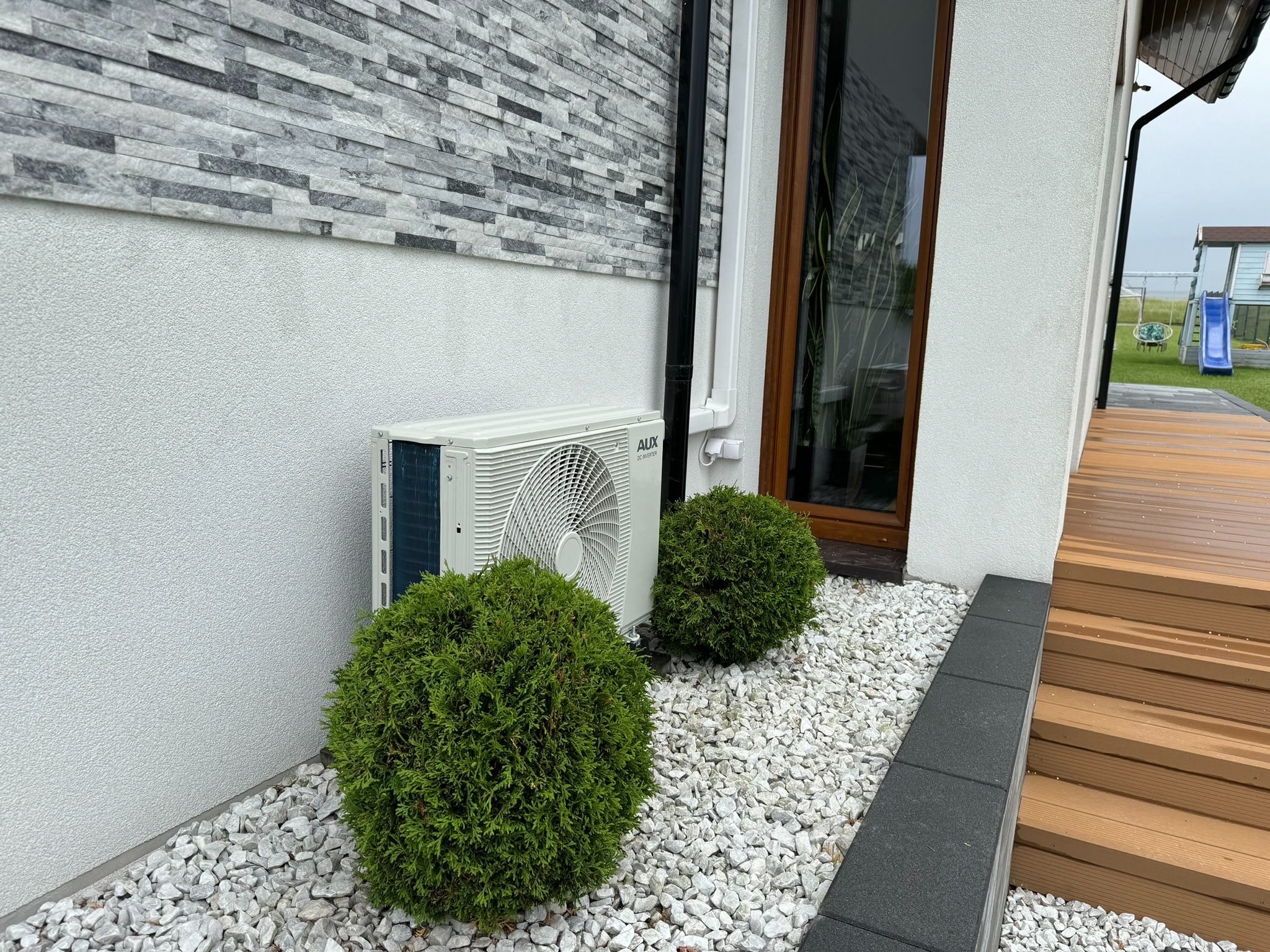 Biała jednostka zewnętrzna klimatyzatora marki AUX zamontowana przy ścianie domu z szarą okładziną, otoczona ozdobnymi krzewami i białym kamieniem, widoczny fragment drewnianego tarasu.
