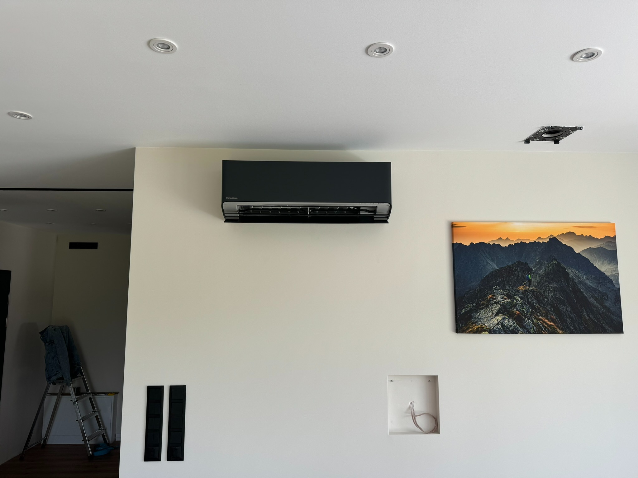 Czarny klimatyzator ścienny marki Panasonic zamontowany w nowoczesnym wnętrzu, obok wisi obraz z górskim krajobrazem, widoczna drabina w tle.