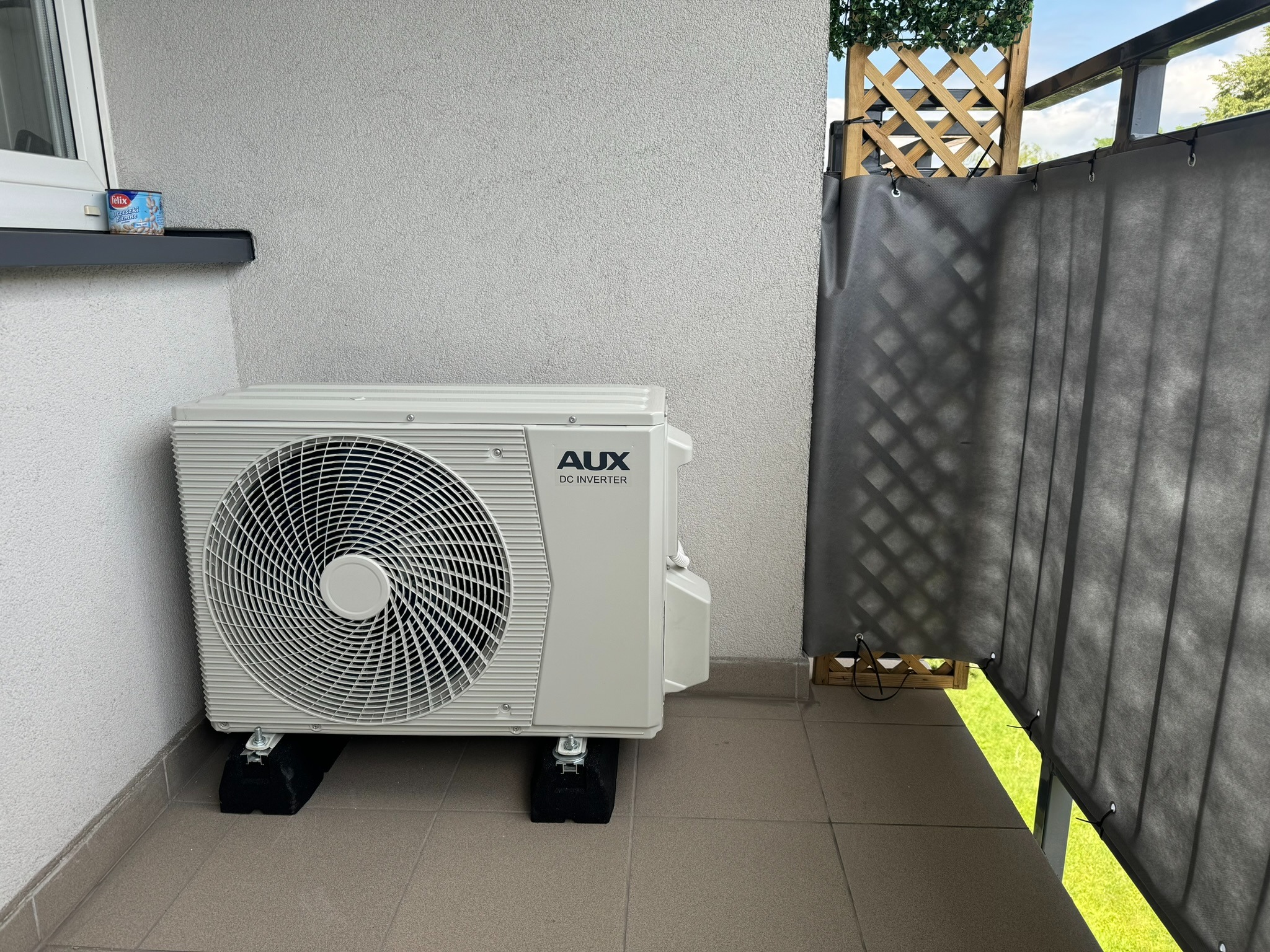 Zewnętrzna jednostka klimatyzacji AUX DC Inverter na balkonie z szarymi płytkami, widoczna puszka jedzenia dla kota i osłona balkonowa