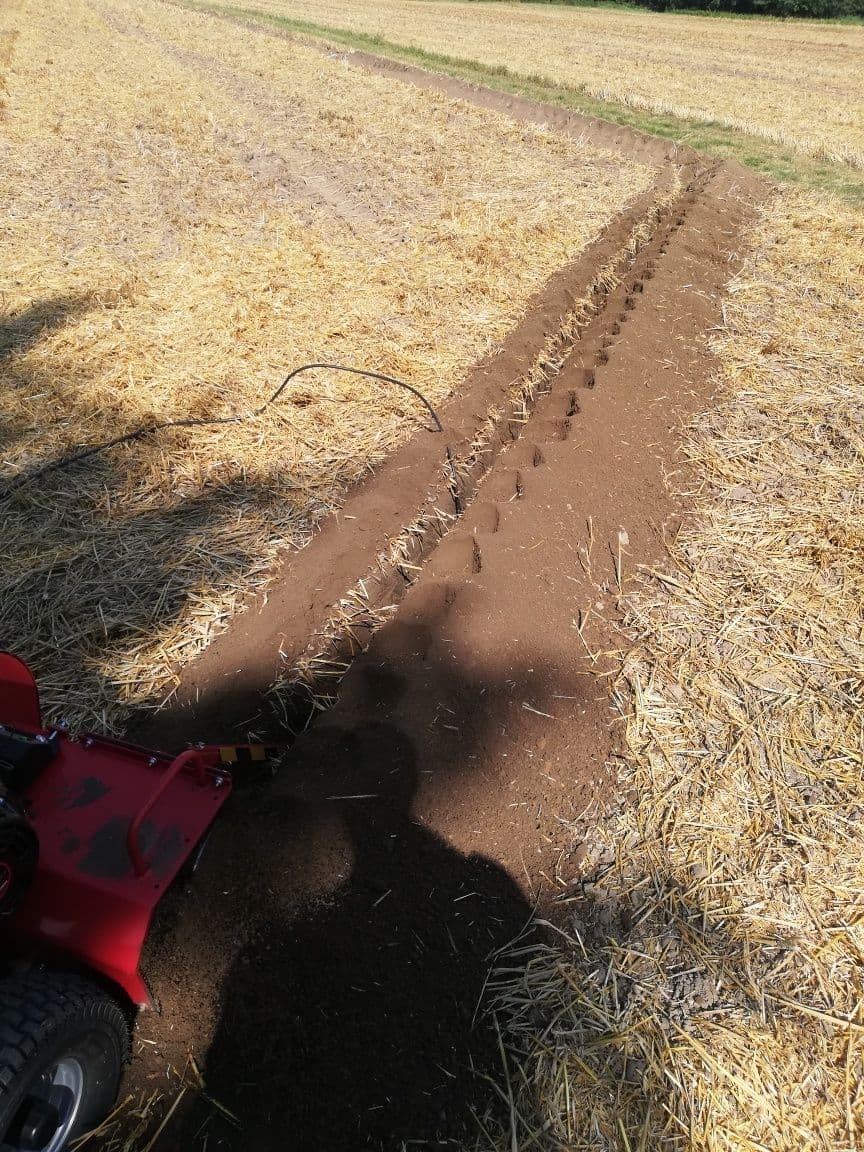 Wąski wykopany rów w polu ze słomą, wykonany za pomocą glebogryzarki z widocznym fragmentem maszyny i cieniem operatora.