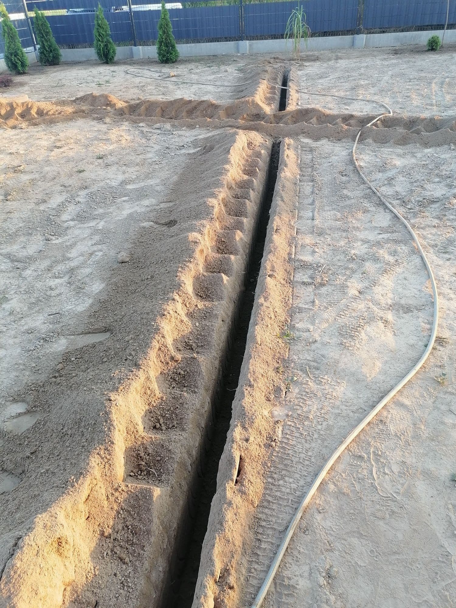 Długi, wąski wykop w piaszczystej ziemi, z widocznymi śladami opon i wystającymi schodkowymi uskokami w ścianach, obok leży zwinięty wąż ogrodowy, w tle rząd tuj i ciemne ogrodzenie.