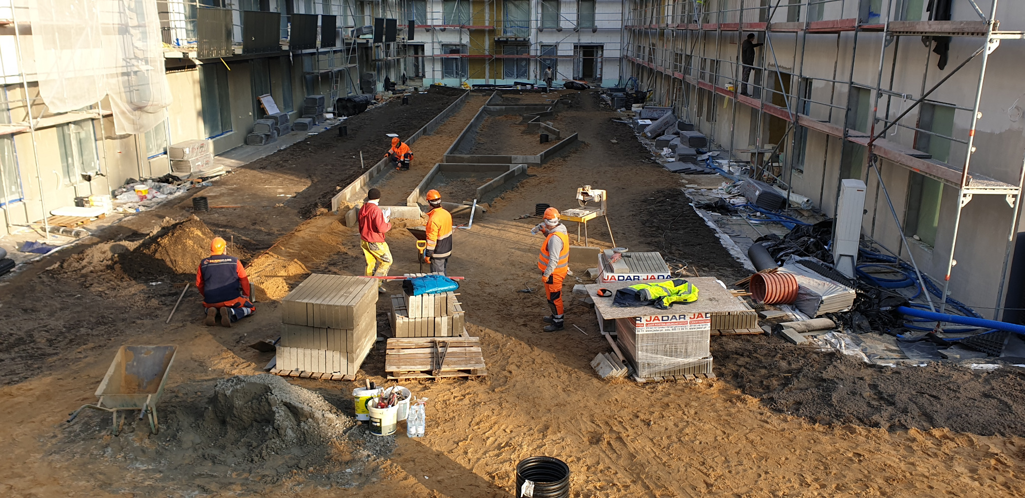 Prace przygotowawcze do układania kostki brukowej na terenie budowy z pracownikami w odblaskowych ubraniach, widoczne krawężniki, kostka brukowa na paletach i rusztowania przy budynku.