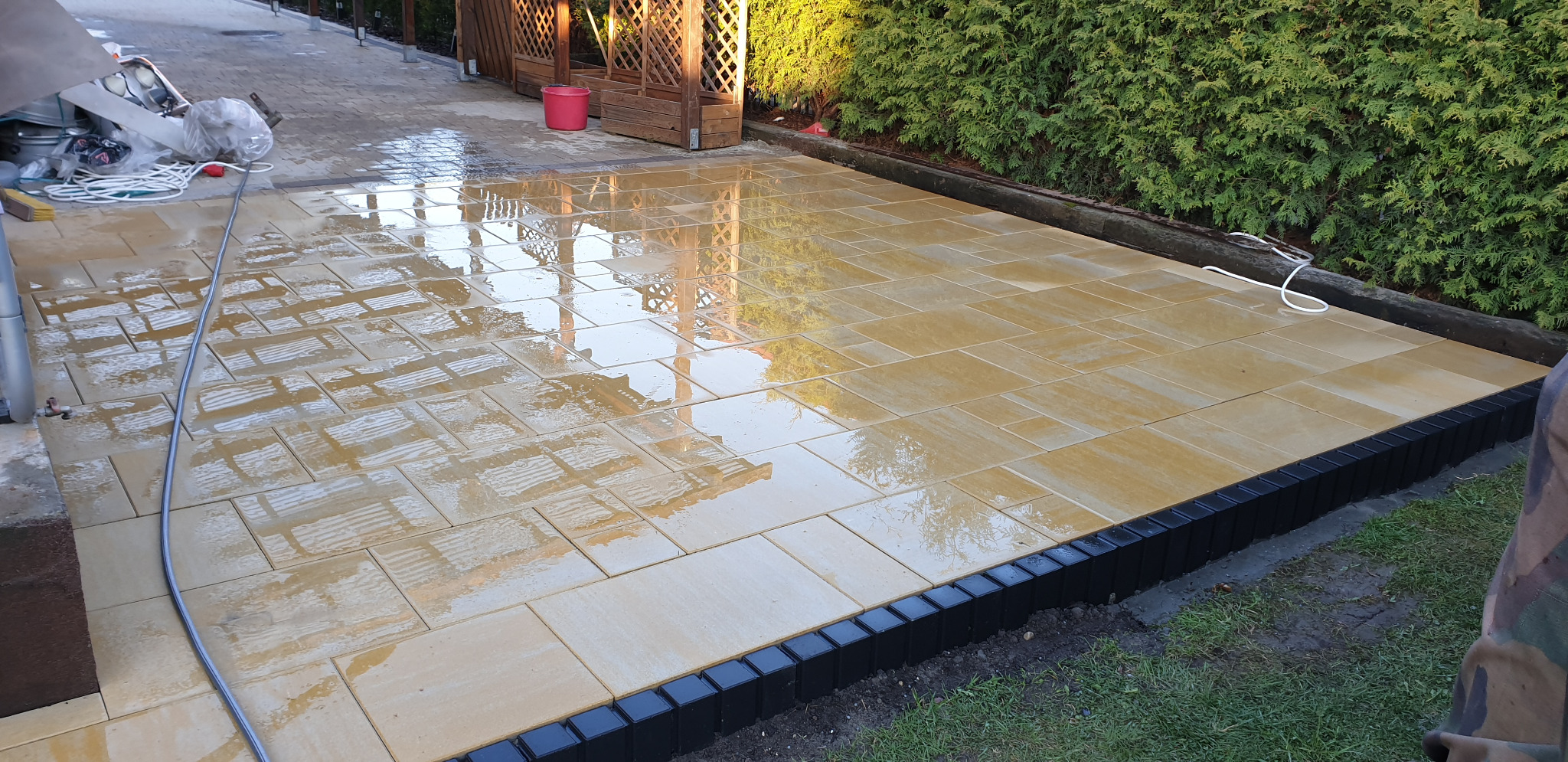 Świeżo ułożona, mokra, beżowa kostka brukowa na posesji, z czarnym obrzeżem, odbijająca niebo i elementy otoczenia, w tle zielona roślinność i drewniana konstrukcja.