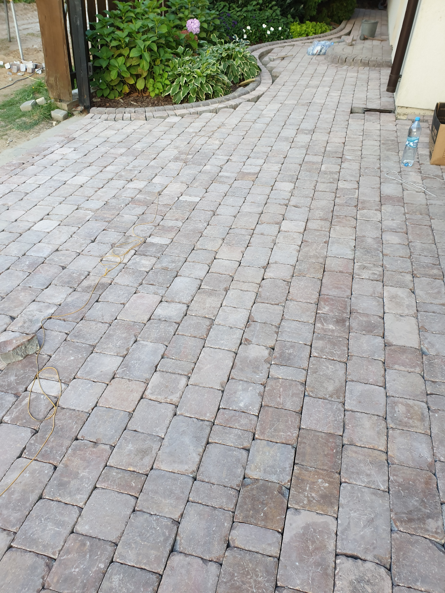 Świeżo ułożona, brązowo-szara kostka brukowa tworząca krętą ścieżkę w ogrodzie, z widocznym fragmentem rabaty kwiatowej i butelką wody mineralnej w tle.