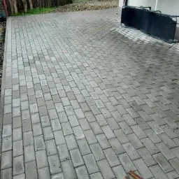 Szara, prostokątna kostka brukowa ułożona na posesji, widoczne dwa drewniane elementy, w tle ogrodzenie i leżak.