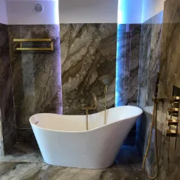 Nowoczesna łazienka z wolnostojącą białą wanną, złotą armaturą, marmurowymi płytkami i podświetleniem LED w kolorze niebieskim.