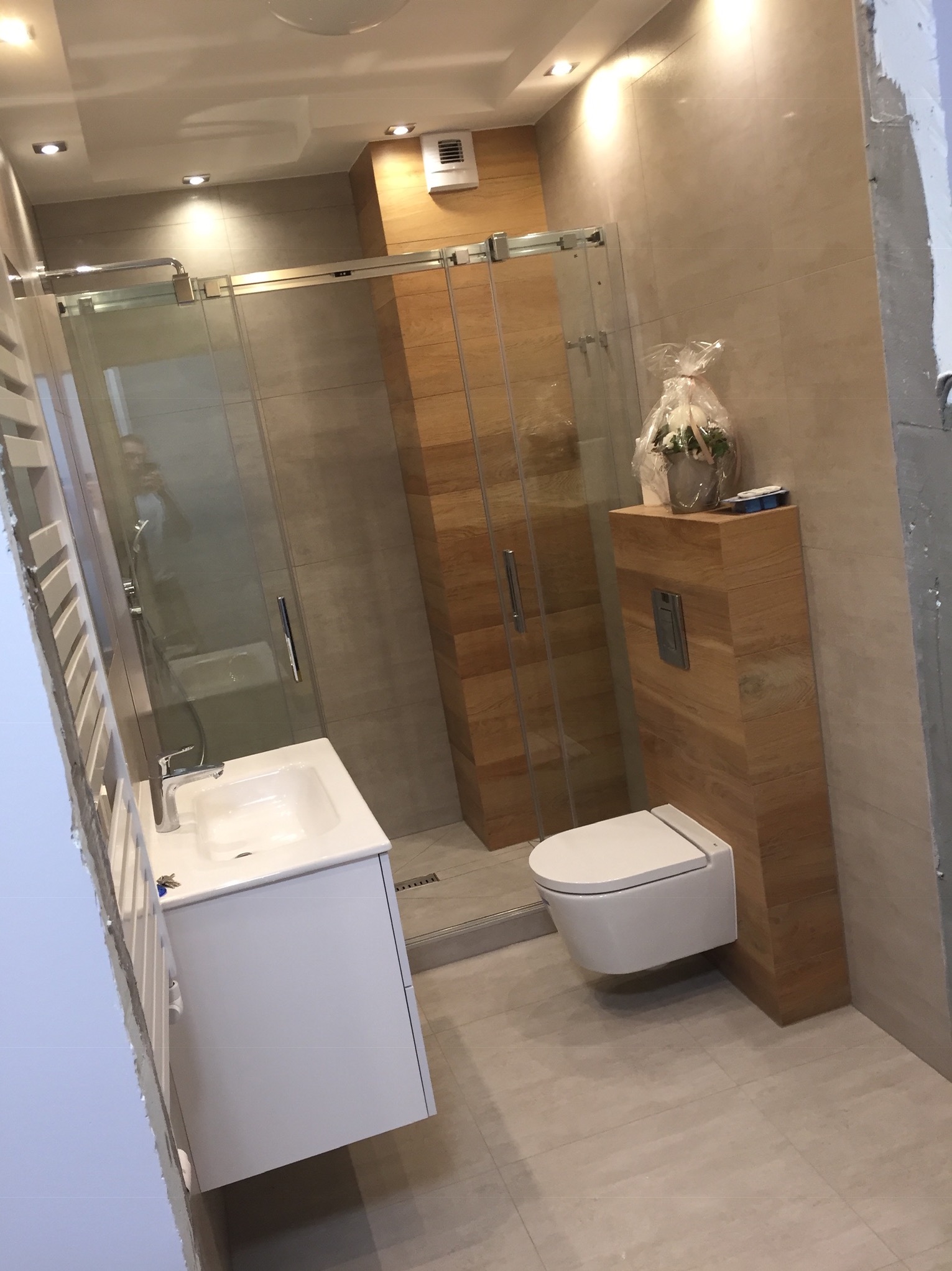 Nowoczesna łazienka w Gdańsku z kabiną prysznicową ze szklanymi drzwiami przesuwnymi, podwieszaną toaletą i umywalką nablatową na białej szafce, wykończona płytkami imitującymi drewno.