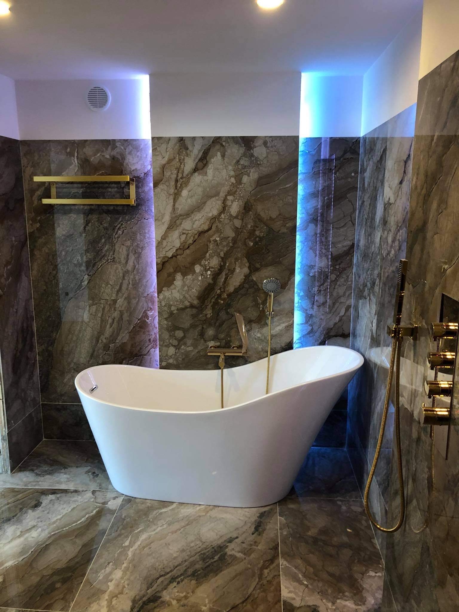 Nowoczesna łazienka z wolnostojącą białą wanną, złotą armaturą, marmurowymi płytkami i podświetleniem LED w kolorze niebieskim.