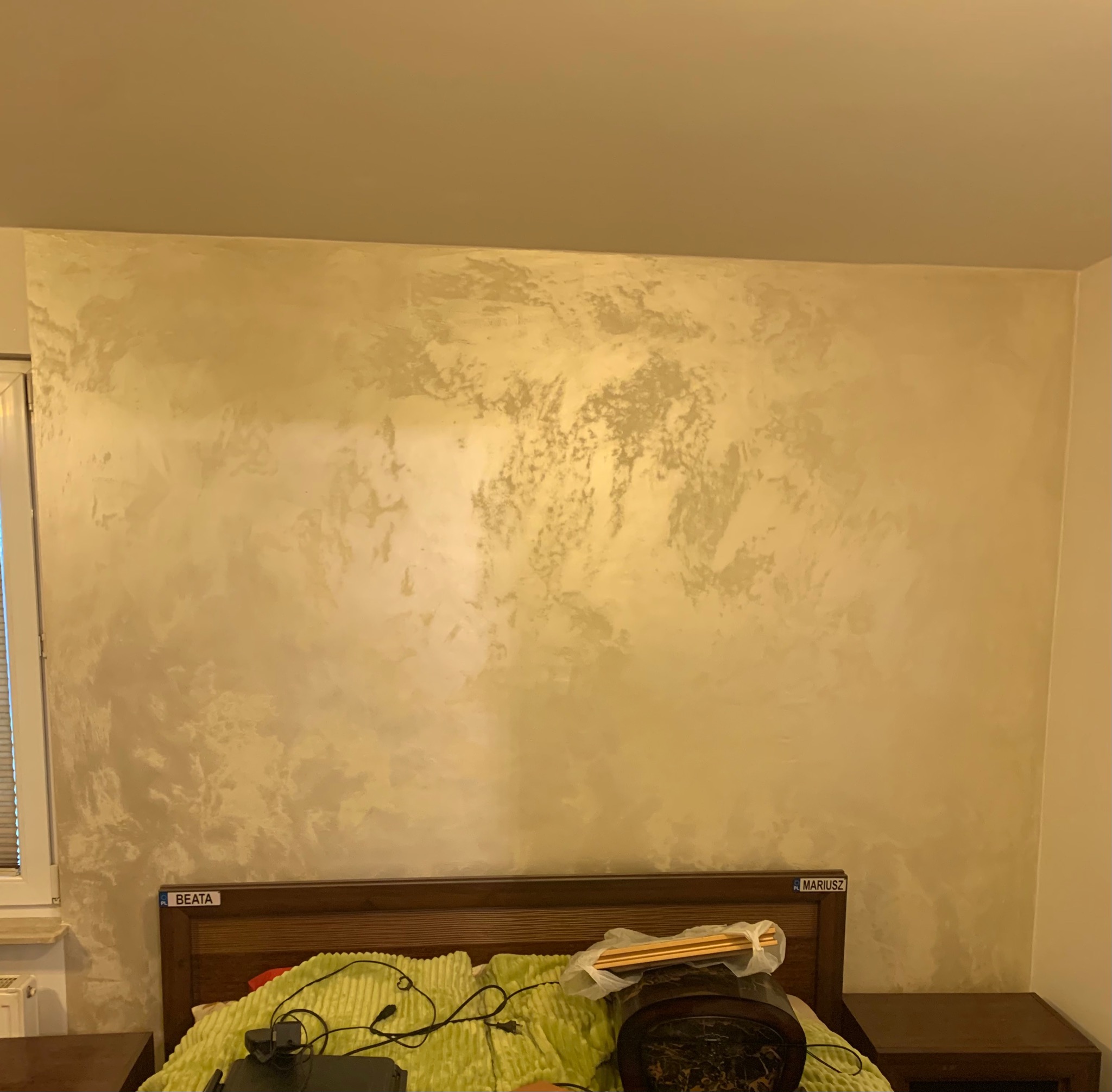 Ściana z efektem dekoracyjnym w odcieniach beżu i srebra, imitująca stiuk wenecki, widoczna nad zagłówkiem łóżka z tabliczkami 'Beata' i 'Mariusz'.
