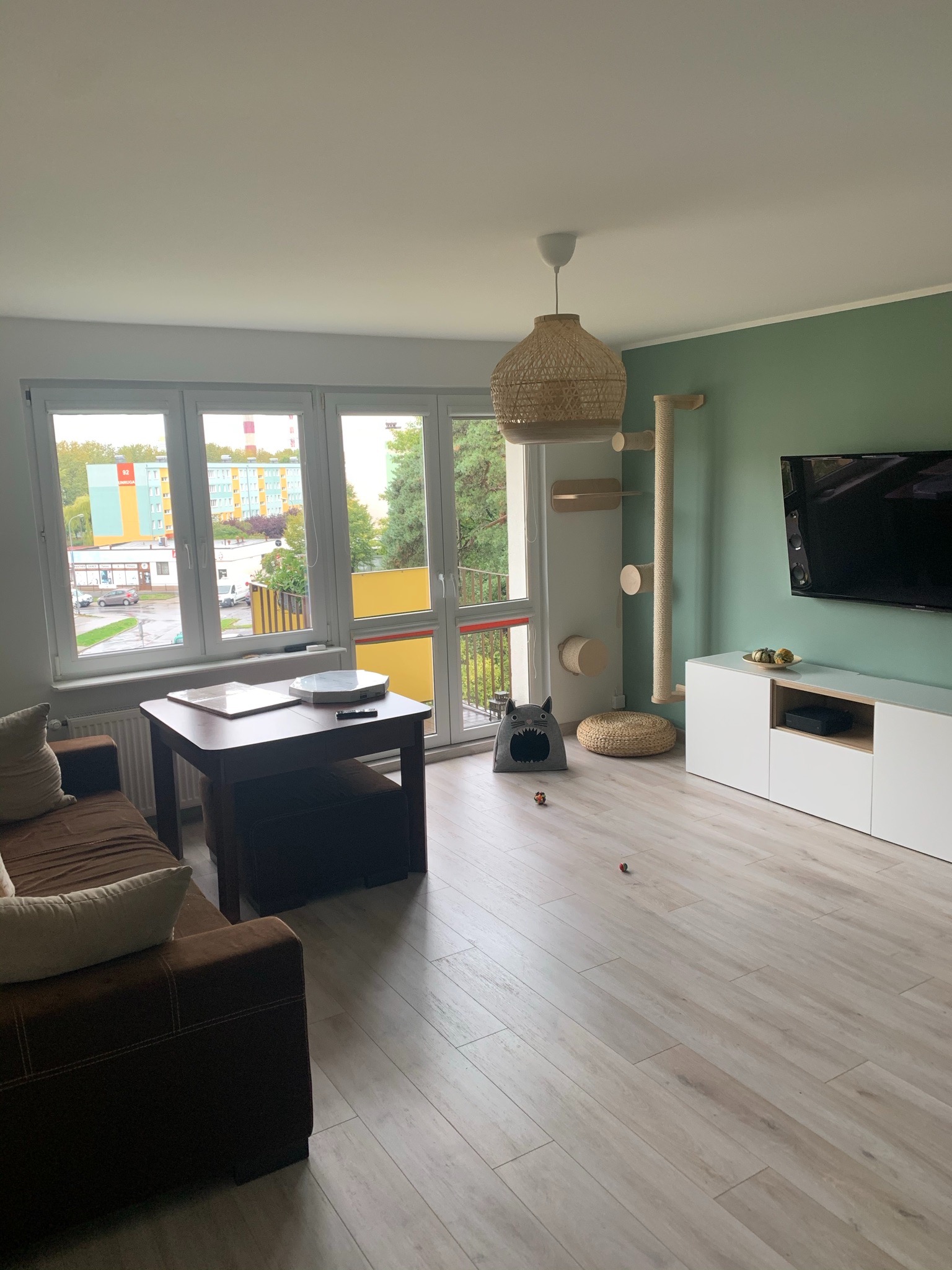 Jasne wnętrze salonu z panelami podłogowymi w odcieniach beżu, brązową sofą, stolikiem kawowym, białą szafką RTV, zieloną ścianą, lampą z wikliny i drapakiem dla kota