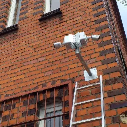 Zewnętrzny montaż trzech kamer przemysłowych na elewacji budynku z czerwonej cegły. Widoczna drabina oparta o ścianę, sugerująca trwający proces instalacji.
