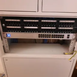 Szafa rack z patch panelem i switchem. Widoczne kable sieciowe, oznaczenia portów i diody sygnalizacyjne. Uziemienie podłączone do obudowy. Biała szafa w pomieszczeniu.