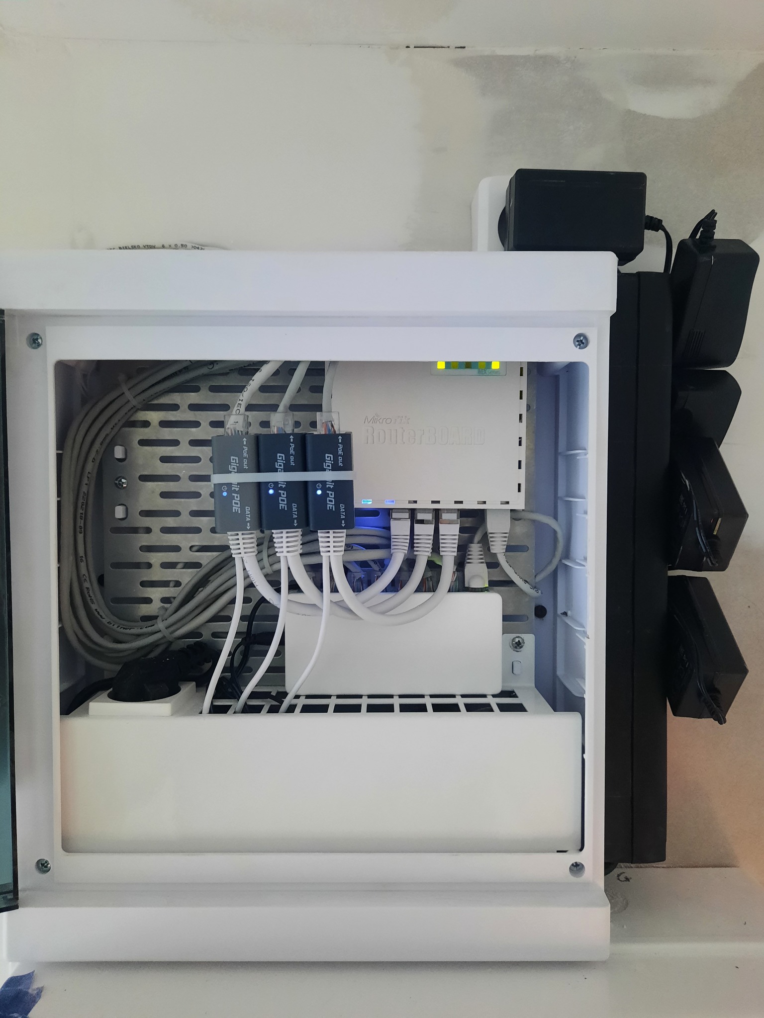Szafka z routerem MikroTik i adapterami Gigabit PoE, okablowanie wewnątrz, zasilacze na zewnątrz. Instalacja sieciowa w białej obudowie, widoczne diody sygnalizacyjne.