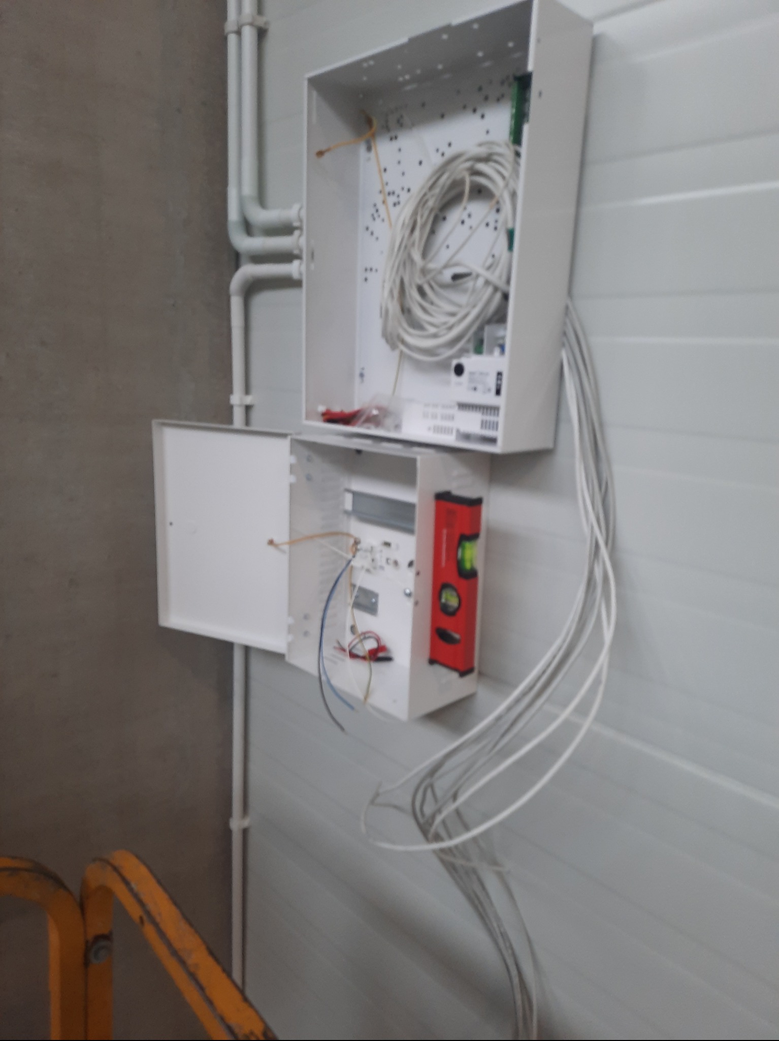Otwarta skrzynka z kablami na ścianie, obok pionowo przymocowana poziomica. Widoczne rury instalacyjne. Elementy instalacji elektrycznej wewnątrz skrzynki.
