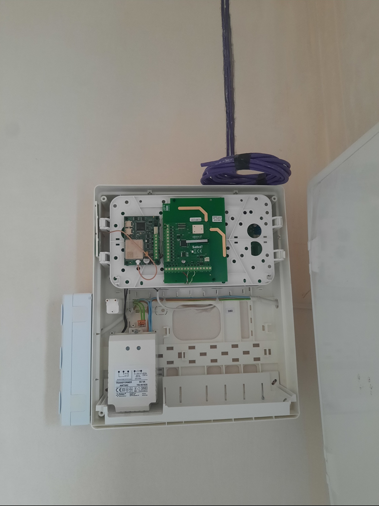 Otwarta, biała skrzynka z elektroniką alarmową, wewnątrz widoczne płytki drukowane i transformator. Nad skrzynką zwój fioletowych kabli. Widok z bliska, na jasnym tle.