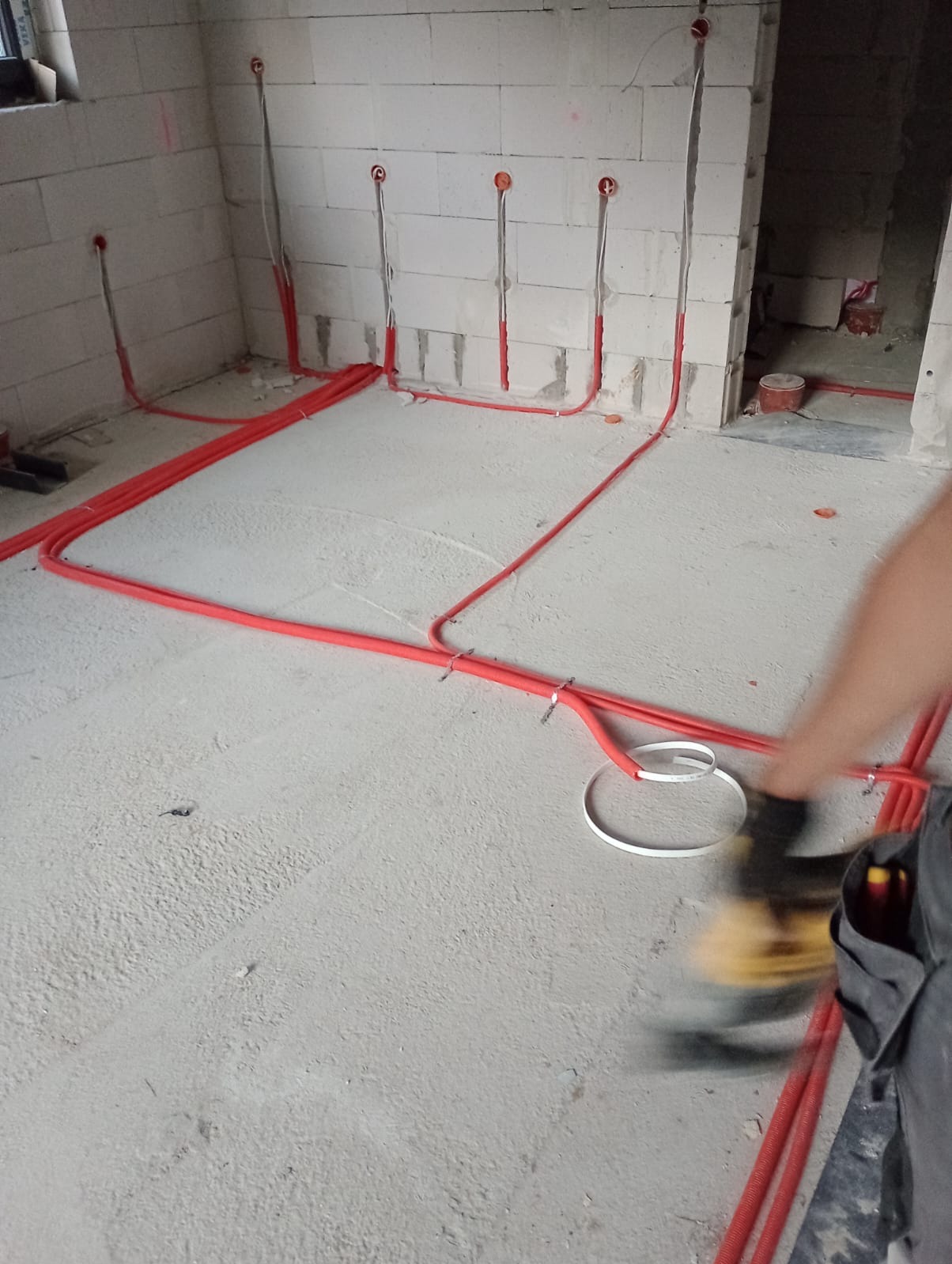 Czerwone rurki instalacji elektrycznej ułożone na surowym betonie w pomieszczeniu z białego bloczka. Widoczne puszki podtynkowe i fragment nogi instalatora.
