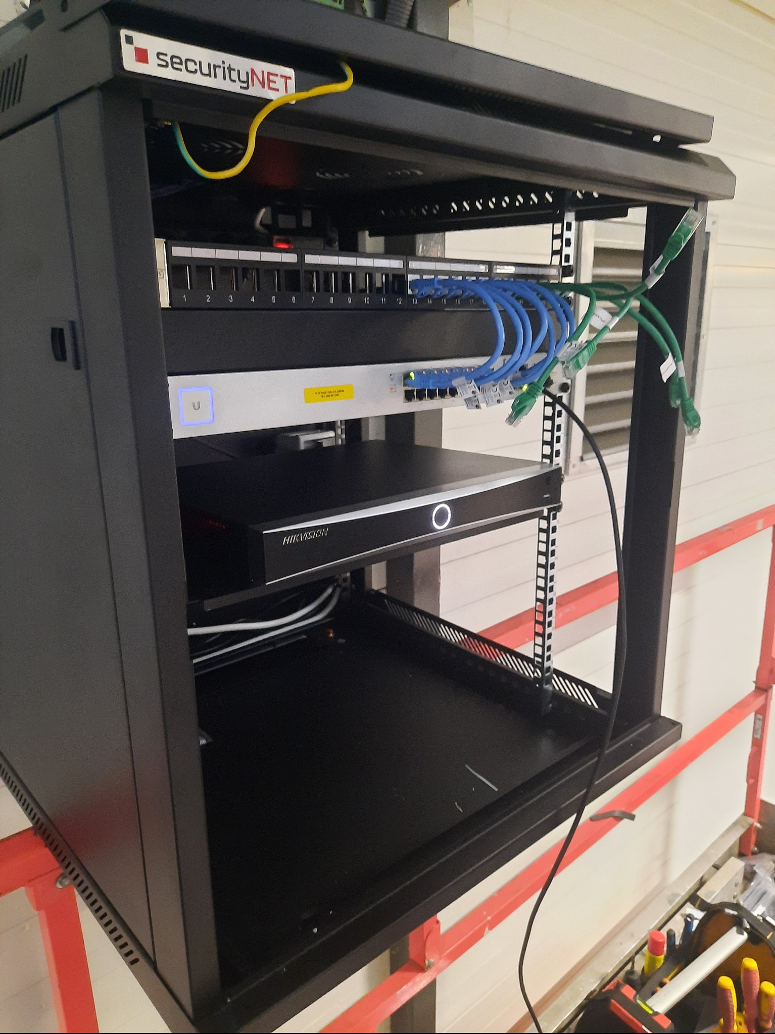 Czarna szafa rack z zamontowanym rejestratorem Hikvision, switchem i patch panelem. Widoczne kable sieciowe i zasilające. Na górze logo SecurityNET. Na dole narzędzia.