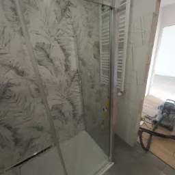 Kabina prysznicowa z wzorem liści na ścianie, zamontowany grzejnik drabinkowy, widoczny odkurzacz przemysłowy i poziomica, pomieszczenie w trakcie prac wykończeniowych.