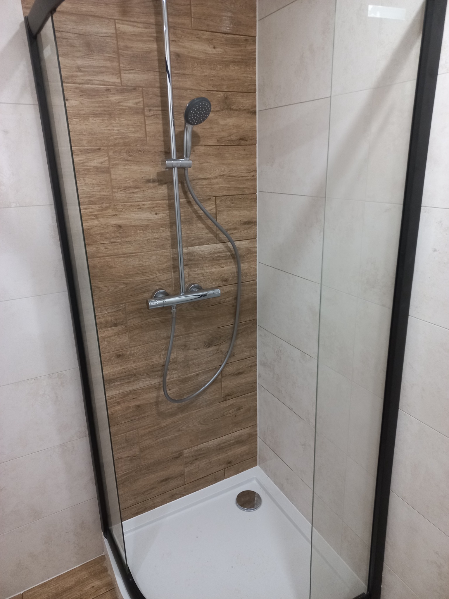 Kabina prysznicowa z czarnymi profilami, wykończona płytkami imitującymi drewno i jasnymi płytkami ceramicznymi, z widoczną baterią termostatyczną i deszczownicą.