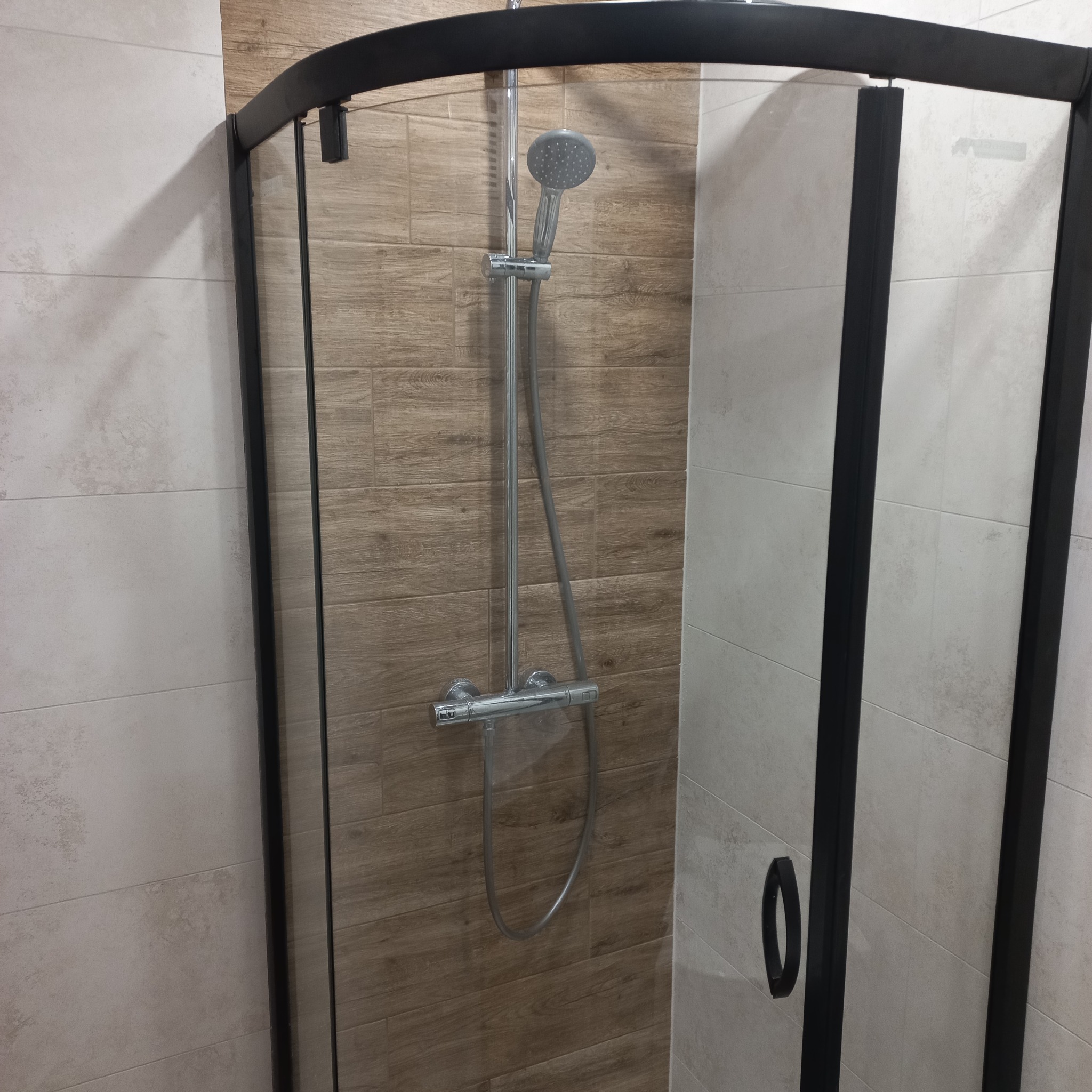 Kabina prysznicowa z czarnymi profilami i deszczownicą zamontowana na ścianie wyłożonej płytkami imitującymi drewno, widoczna część ściany z jasnymi płytkami.