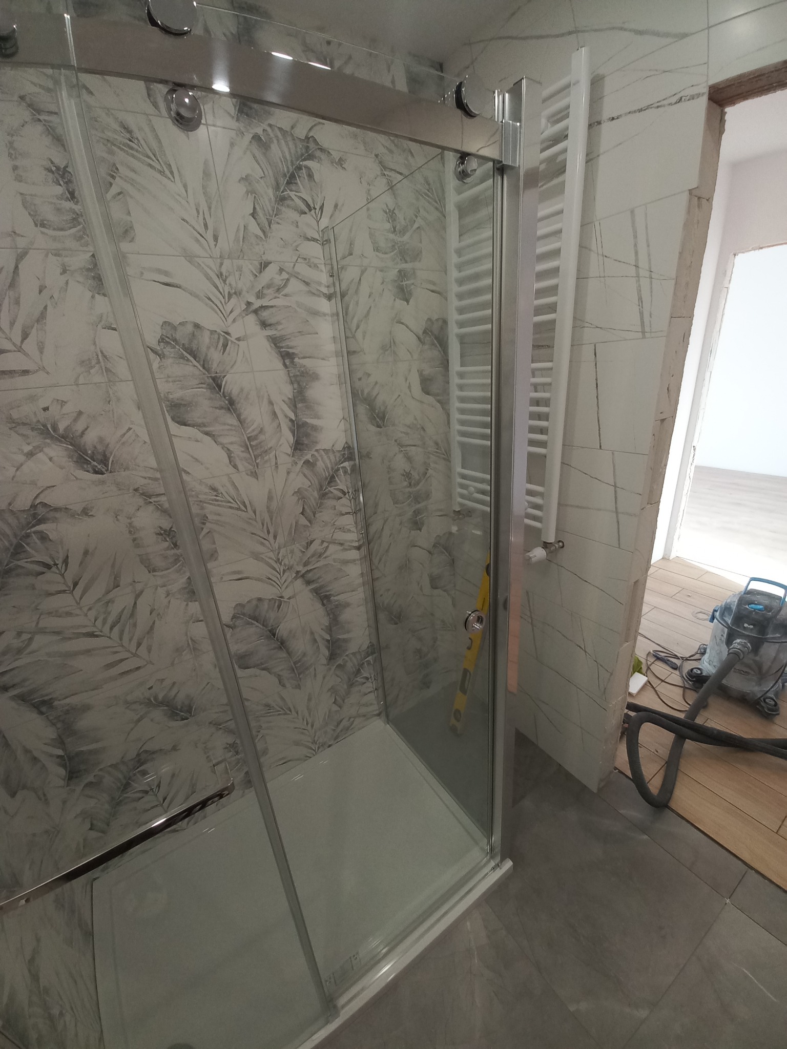 Kabina prysznicowa z wzorem liści na ścianie, zamontowany grzejnik drabinkowy, widoczny odkurzacz przemysłowy i poziomica, pomieszczenie w trakcie prac wykończeniowych.