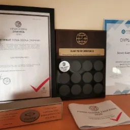 Zestaw certyfikatów i dyplomów 'Firma Godna Zaufania' oraz nagród, w tym dyplom dla Doroty Karpińskiej za sprzedaż największej polisy grupowej na życie w Comperia Ubezpieczenia, obok długopis...