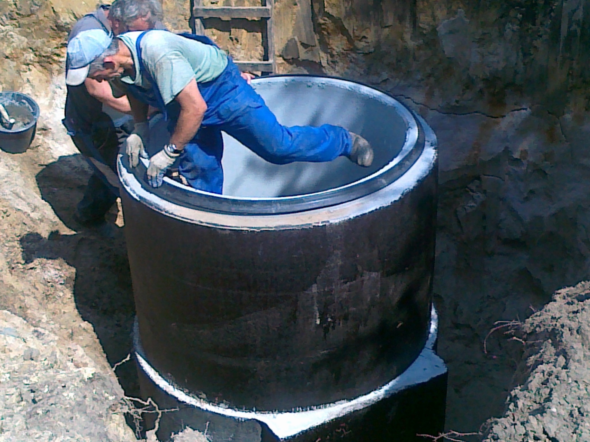 Montaż betonowego kręgu studni w wykopie przez dwóch pracowników, jeden wchodzi do środka dla precyzyjnego ustawienia, widoczny kubeł z zaprawą.