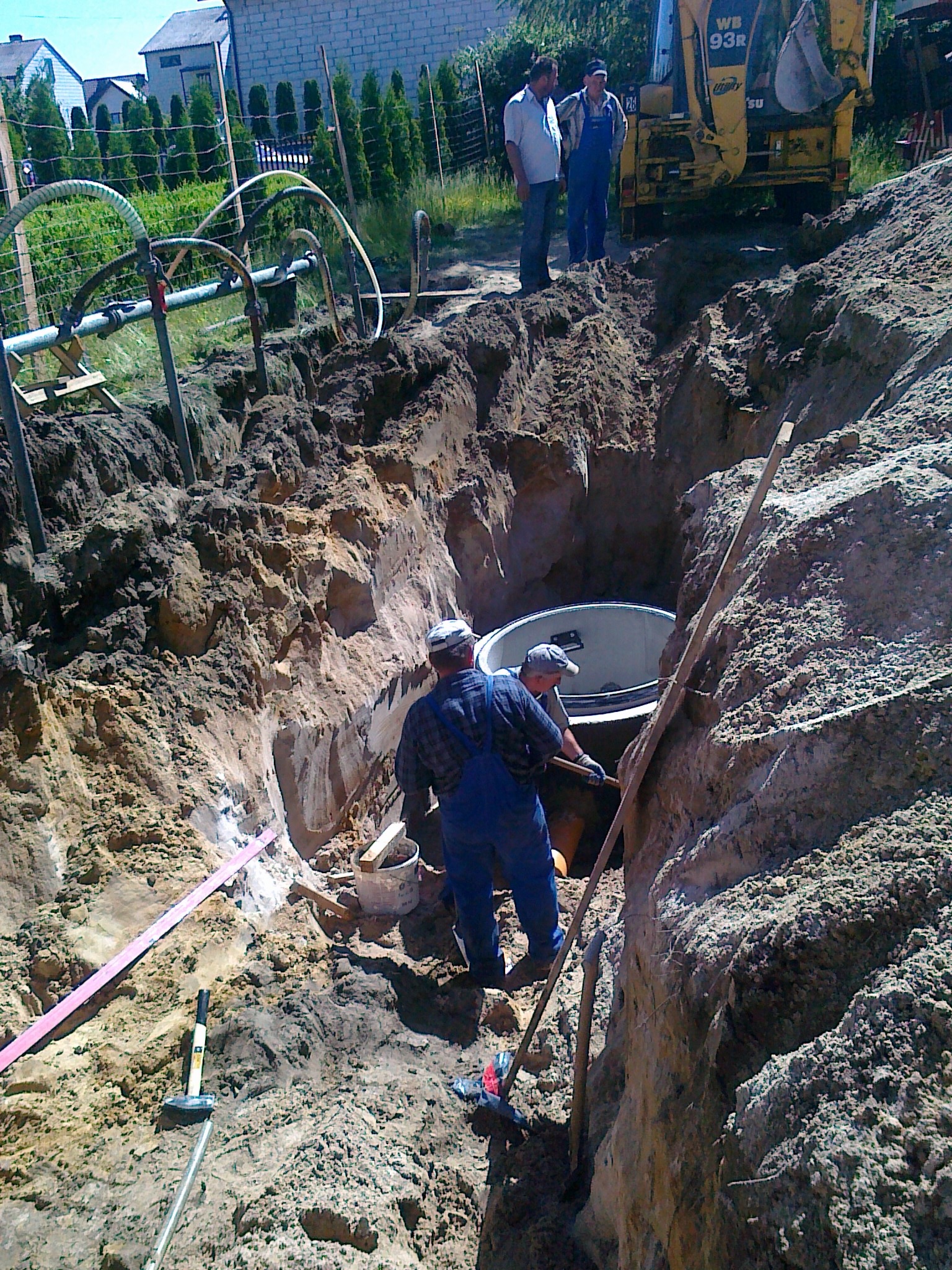 Głęboki wykop w ziemi z betonowym zbiornikiem w środku, dwóch robotników w kombinezonach w dole, łopata oparta o ścianę wykopu, na górze koparka i dwaj obserwujący mężczyźni.