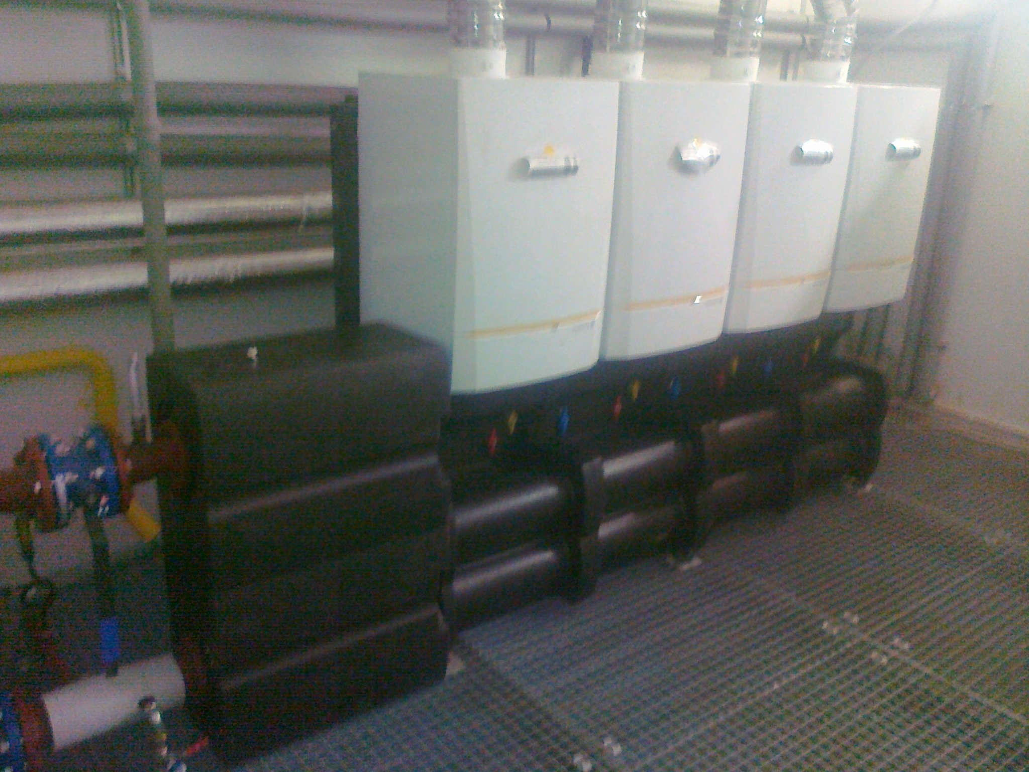 Rząd białych pieców gazowych zamontowanych na czarnej podstawie, widoczne rury i zawory instalacji, całość na metalowej kratownicy podłogowej.