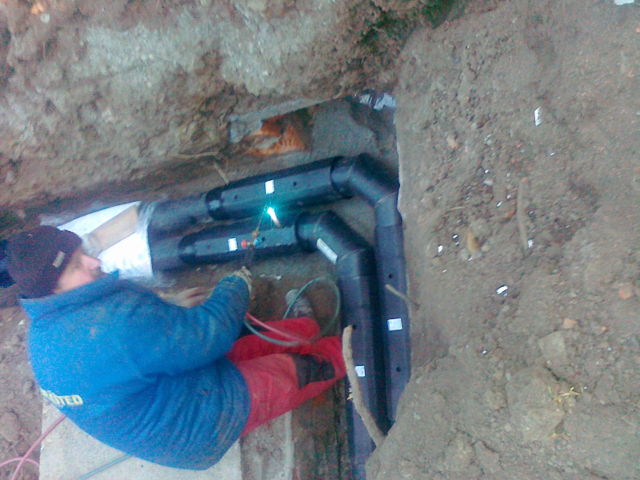 Pracownik w niebieskiej kurtce łączy rury instalacji sanitarnej w wykopie ziemnym, widoczne złącza i kable.