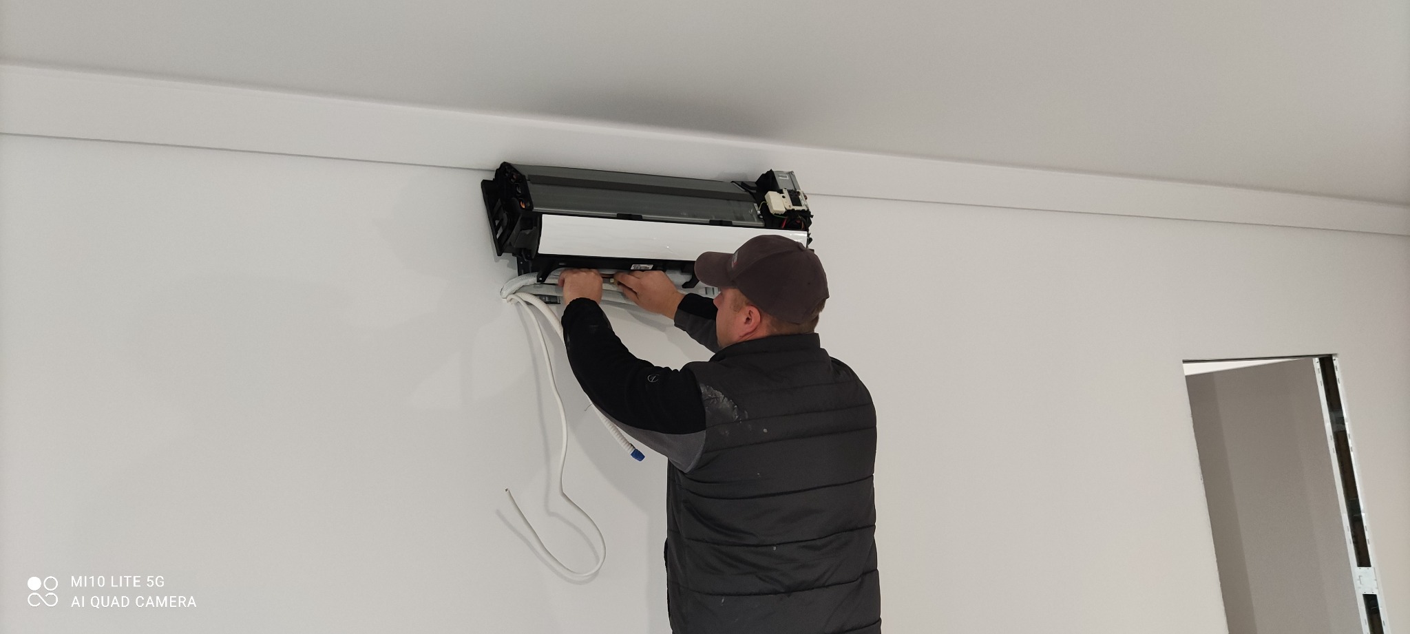 Montaż klimatyzacji w pomieszczeniu o białych ścianach; widoczny pracownik w czapce i kamizelce podłączający przewody do jednostki wewnętrznej klimatyzatora.