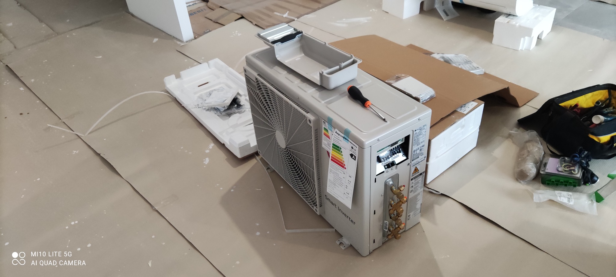 Rozpakowana jednostka zewnętrzna klimatyzatora LG Smart Inverter z naklejką energetyczną A, leżąca na kartonie z wkrętakiem na obudowie, obok torby narzędziowej i opakowania z elementami montażowymi.