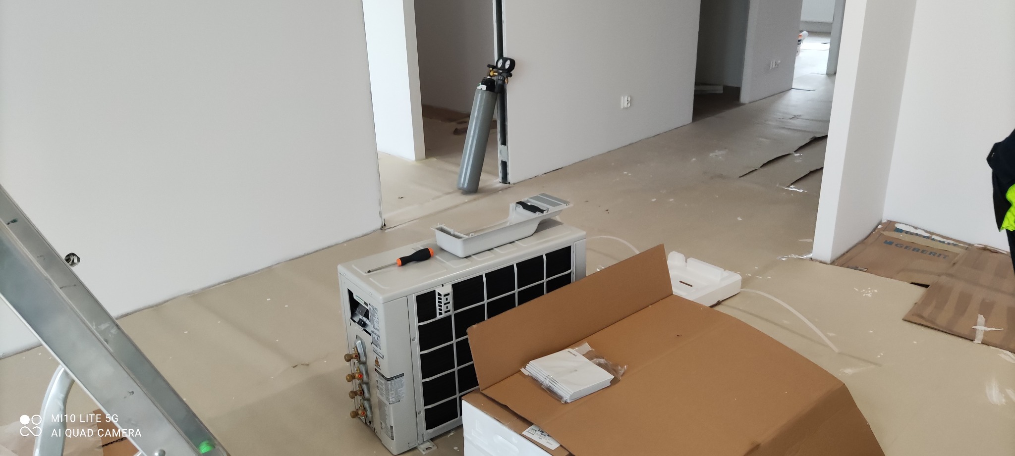 Jednostka zewnętrzna klimatyzacji z osprzętem i butlą z gazem chłodniczym w surowym wnętrzu podczas montażu, widoczna drabina i kartony zabezpieczające podłogę.