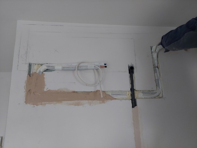 Wnęka w ścianie z instalacją hydrauliczną w trakcie montażu, widoczne rury, kable, pianka montażowa i częściowo nałożona zaprawa.