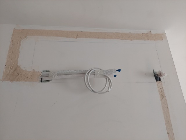 Przygotowanie ściany pod montaż klimatyzacji: wycięty otwór, wystające przewody w osłonie, obramowanie otworu w trakcie szpachlowania.