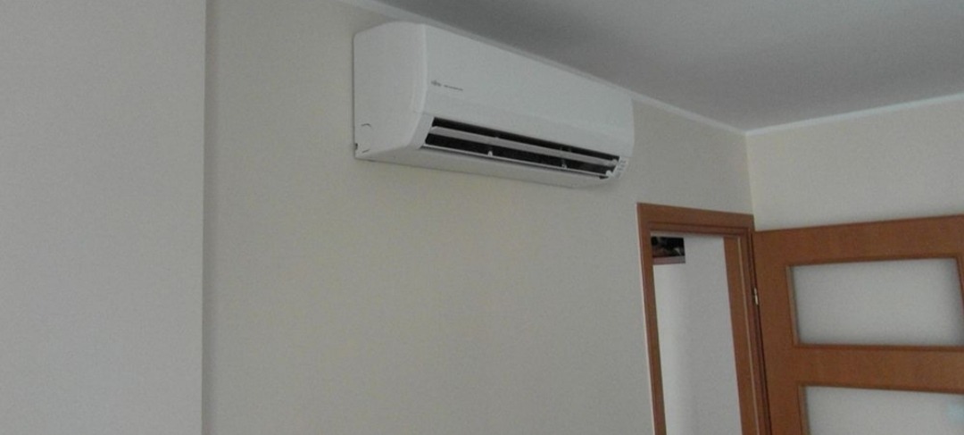 Biały klimatyzator ścienny marki Fujitsu zamontowany w rogu jasnego pokoju z widocznym fragmentem drewnianych drzwi z szybami.