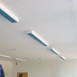 Biały perforowany sufit podwieszany z zamontowanymi, częściowo jeszcze zabezpieczonymi folią, liniowymi lampami LED, widok z dołu na fragment pomieszczenia w trakcie remontu.