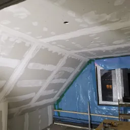 Poddasze w trakcie wykańczania płytami gipsowo-kartonowymi, widoczne łączenia szpachlowane, okno zabezpieczone folią, narzędzia na drewnianym podeście.