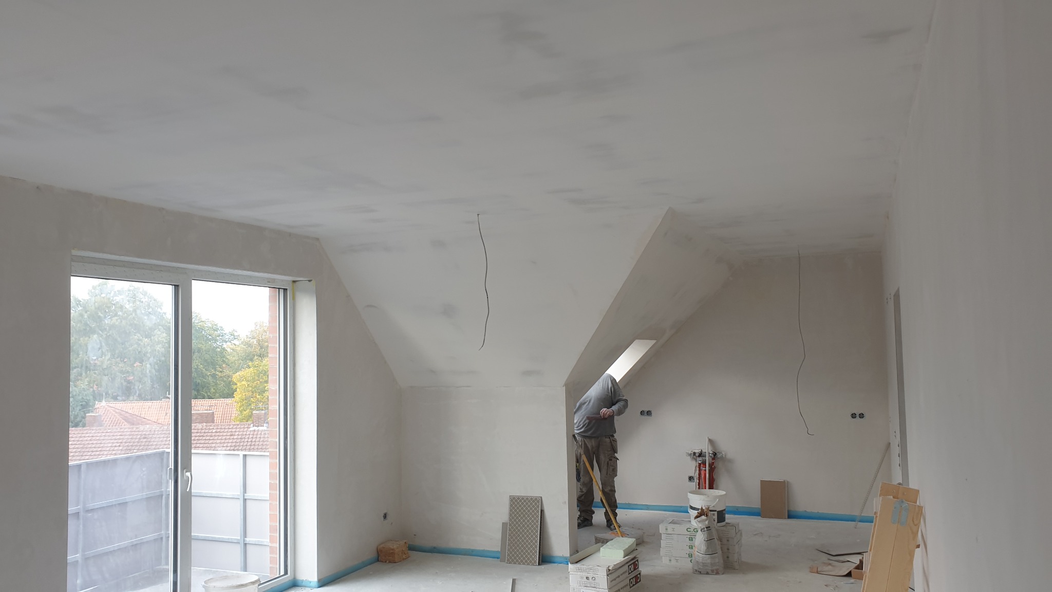 Poddasze w trakcie wykańczania: pracownik szpachluje sufit na mokro, widoczne narzędzia, materiały budowlane i okno balkonowe z widokiem na zewnątrz.