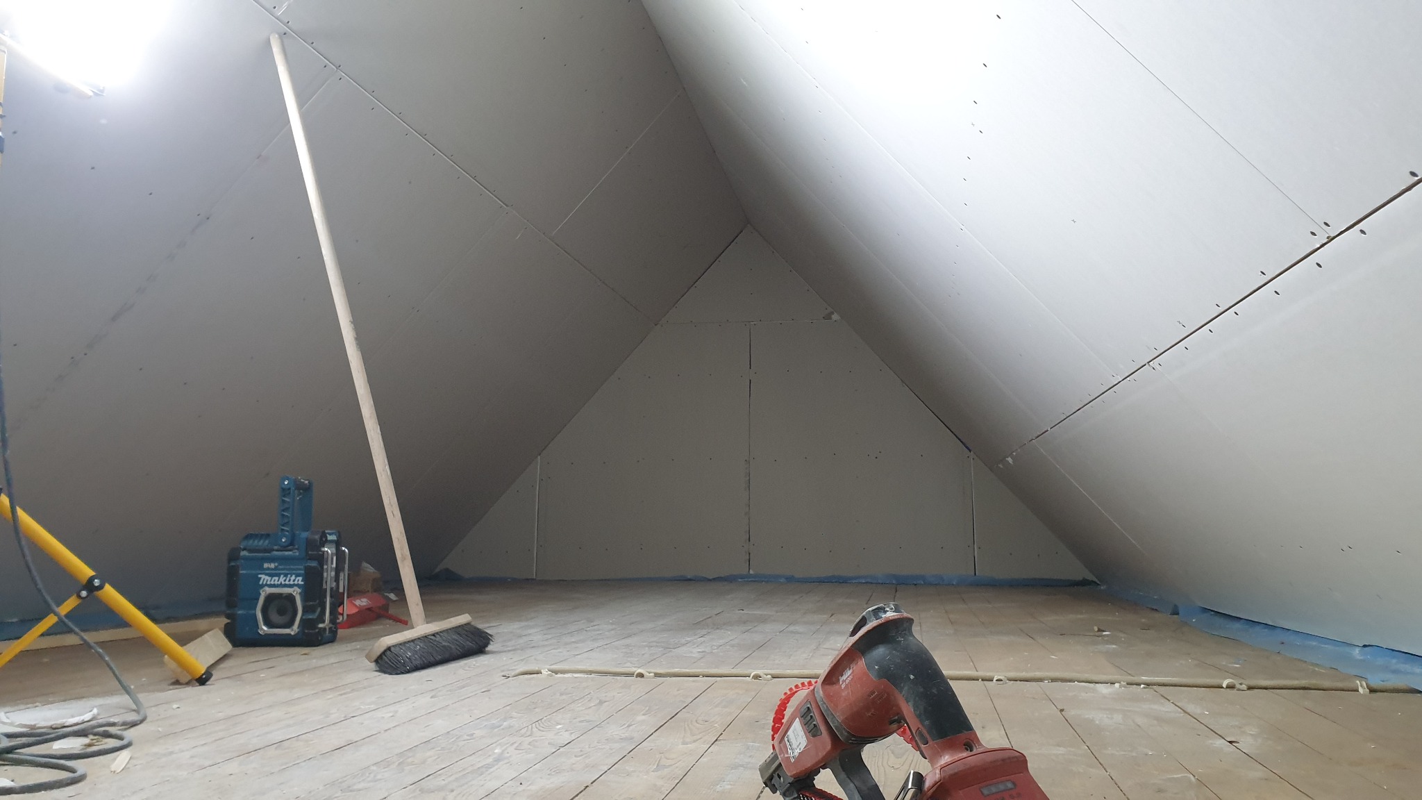 Poddasze w trakcie wykańczania płytami gipsowo-kartonowymi, widoczne mocowania, narzędzia budowlane i lampa oświetleniowa.