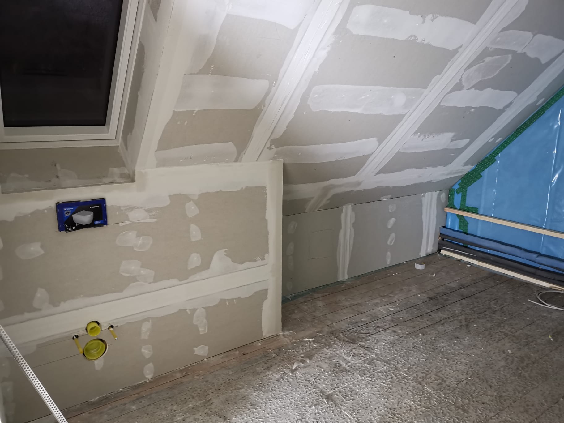 Poddasze w trakcie wykańczania: płyty kartonowo-gipsowe na skosach i ścianach, z widocznymi łączeniami i szpachlowaniem. Instalacja sanitarna z żółtymi rurami wyprowadzona ze ściany. Częściowo...