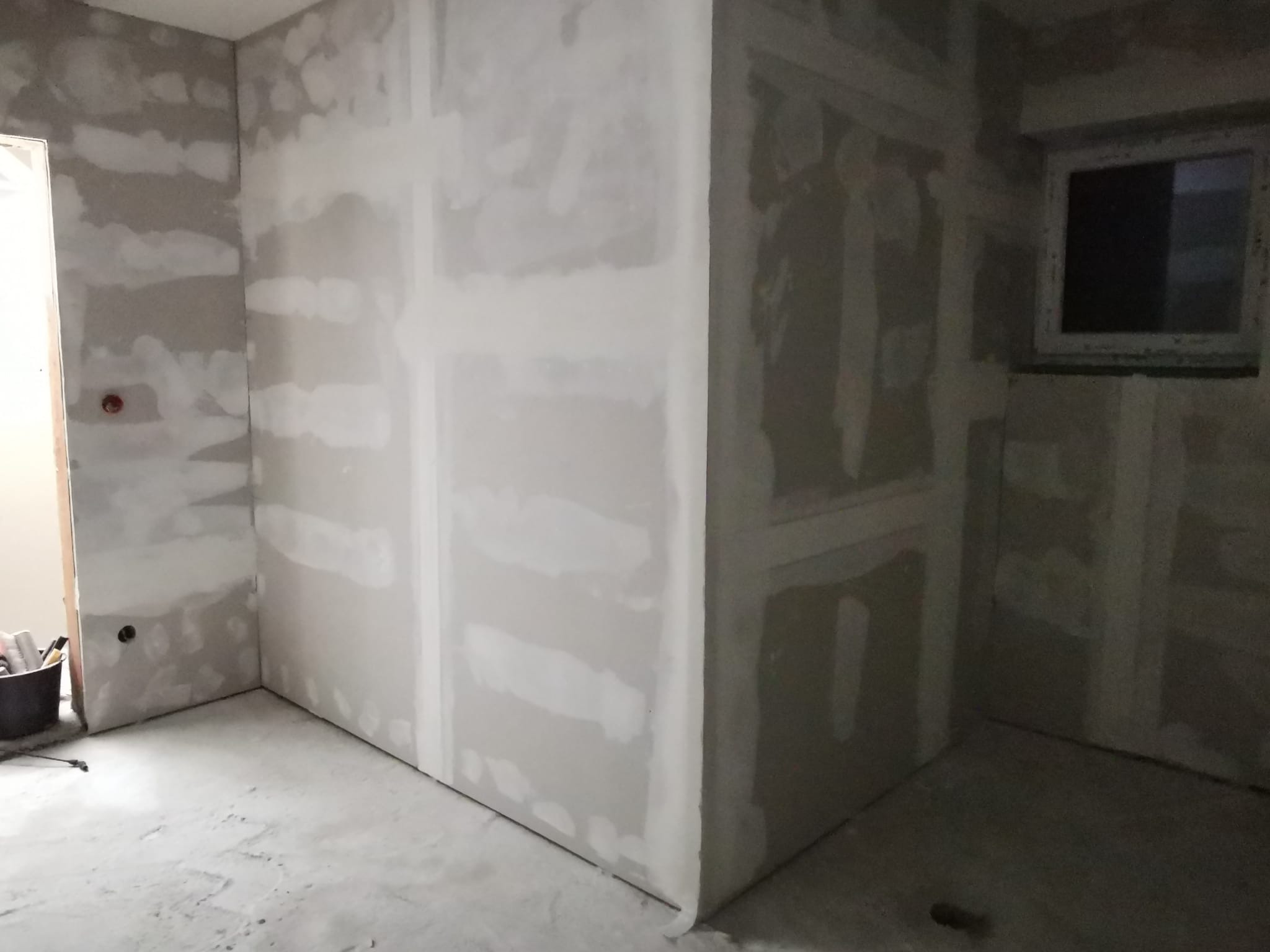 Wnętrze w trakcie remontu, ściany wykończone płytami kartonowo-gipsowymi, widoczne łączenia płyt przed szpachlowaniem, surowy beton na podłodze, małe okno w ścianie.