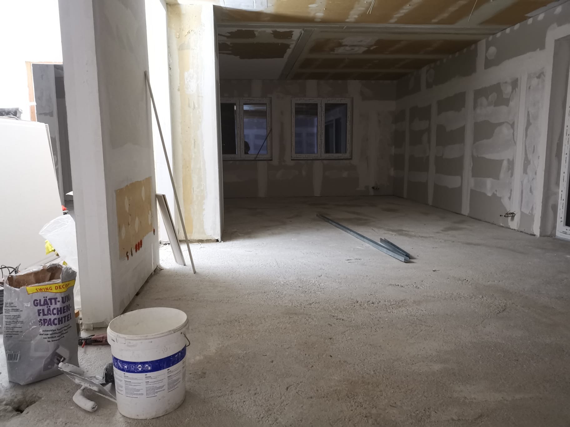 Wnętrze w trakcie remontu: ściany z płyt kartonowo-gipsowych przed szpachlowaniem, widoczne profile metalowe, wiadro z masą szpachlową i worek gładzi, surowy beton na podłodze.