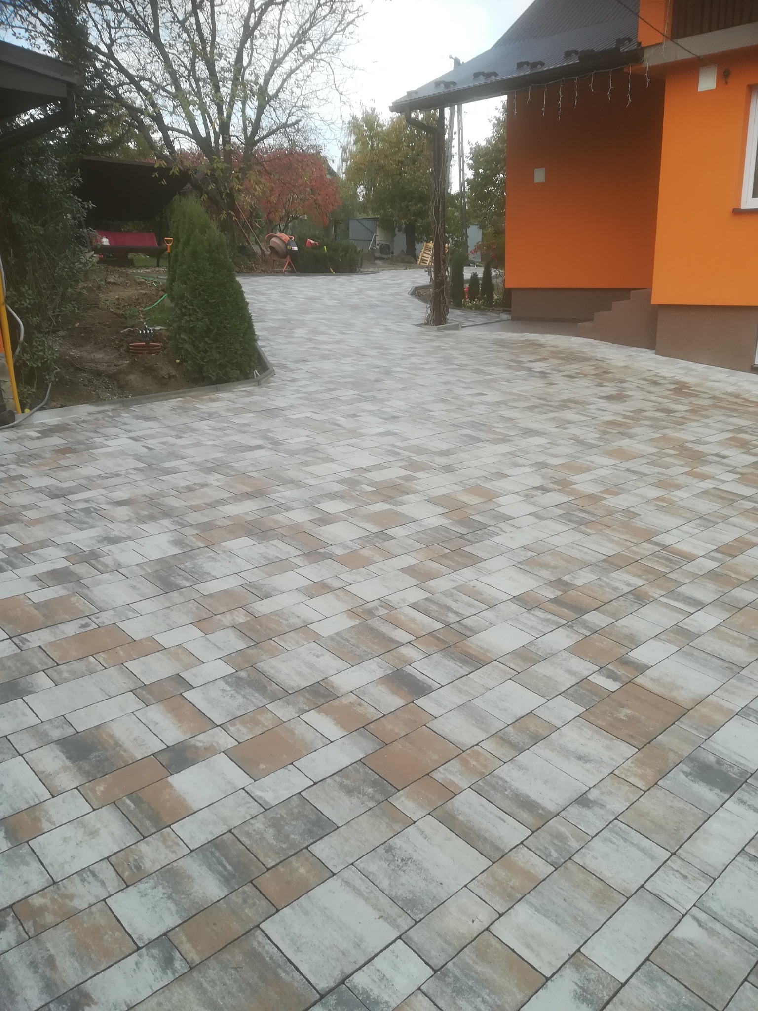 Ułożona kostka brukowa w odcieniach szarości, beżu i brązu prowadząca do pomarańczowego domu z widoczną zielenią w tle.
