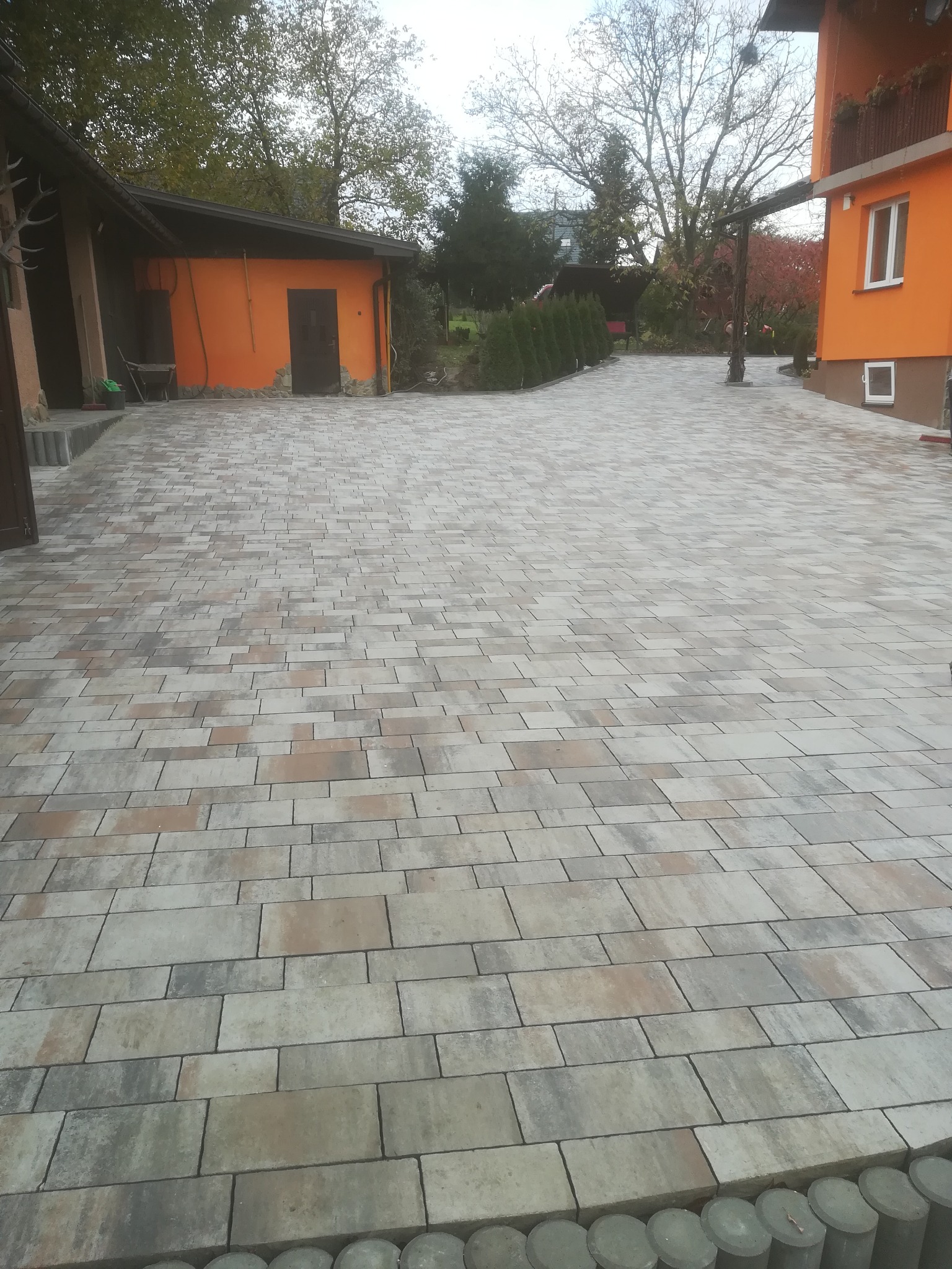 Ułożona nawierzchnia z kostki brukowej w odcieniach szarości i beżu, otoczona budynkami w kolorze pomarańczowym i zielenią drzew.