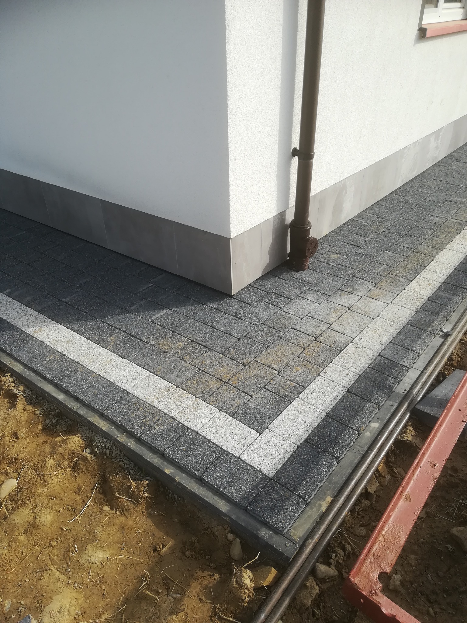Fragment ułożonej szarej kostki brukowej z białym pasem obok ściany budynku z rynną, widoczne narzędzia i ziemia.
