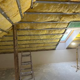 Usługi minikoparką Edengard Andrzej Folwarski - Poddasze w trakcie izolacji termicznej, widoczne żółte ocieplenie między metalowymi profilami, okno dachowe z zieloną osłoną, drewniany słup i drabina, lampa budowlana na statywie.