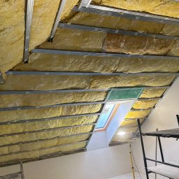 Usługi minikoparką Edengard Andrzej Folwarski - Poddasze w trakcie prac izolacyjnych, widoczna wełna mineralna między metalowymi profilami, okno dachowe z folią ochronną, rusztowanie w tle.