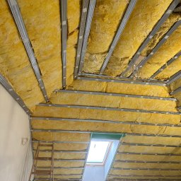 Usługi minikoparką Edengard Andrzej Folwarski - Żółta wełna mineralna izolująca skośny dach poddasza, zamontowana między metalowymi profilami, widoczne okno dachowe i drabina oparta o ścianę.