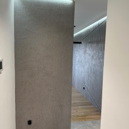Usługi minikoparką Edengard Andrzej Folwarski - Nowoczesny korytarz z dekoracyjną ścianą pokrytą szarą, strukturalną gładzią, oświetleniem LED i podłogą z płytek imitujących drewno.